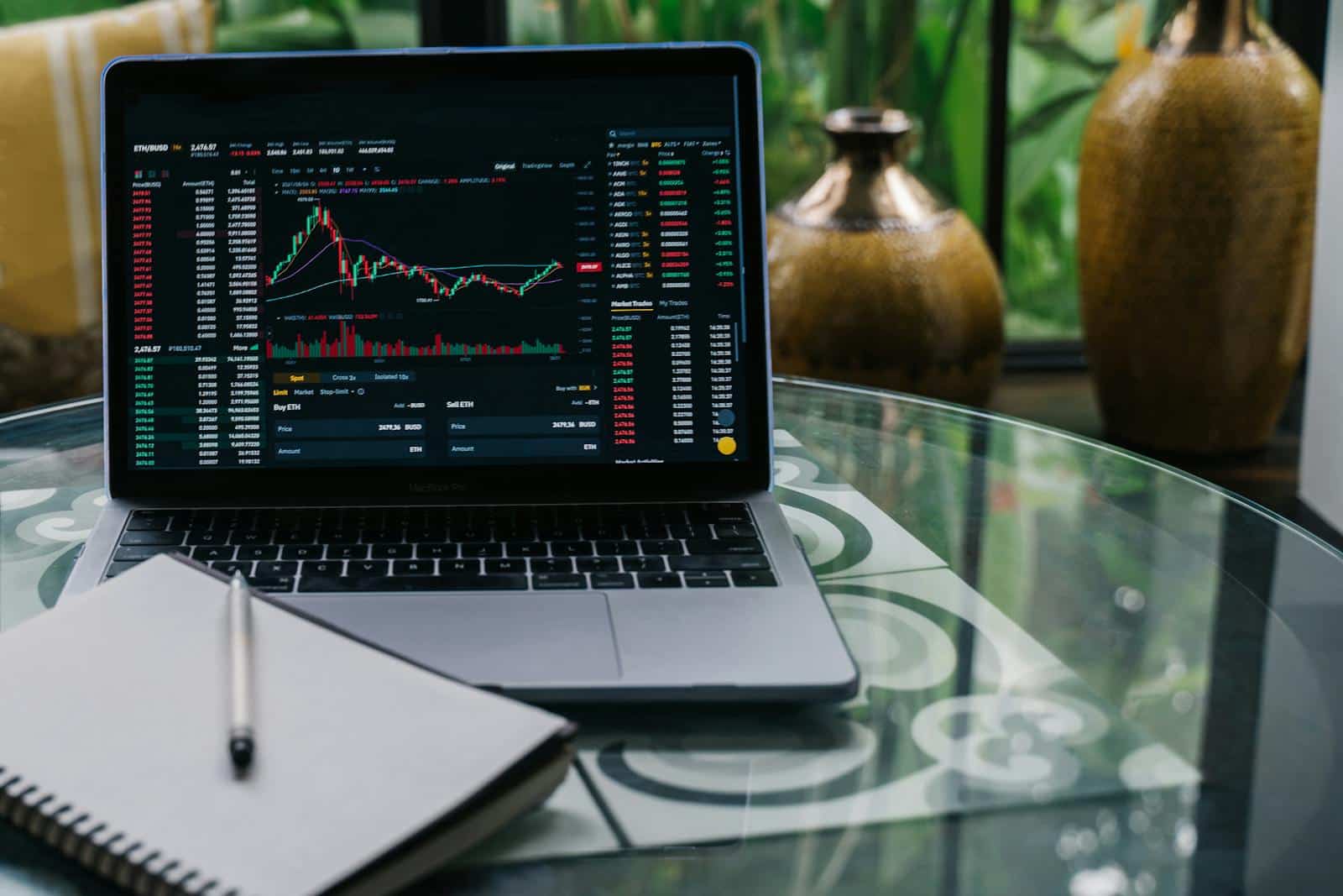 Laptop displaying cryptocurrency stocks and graphs on a glass table with a notepad.