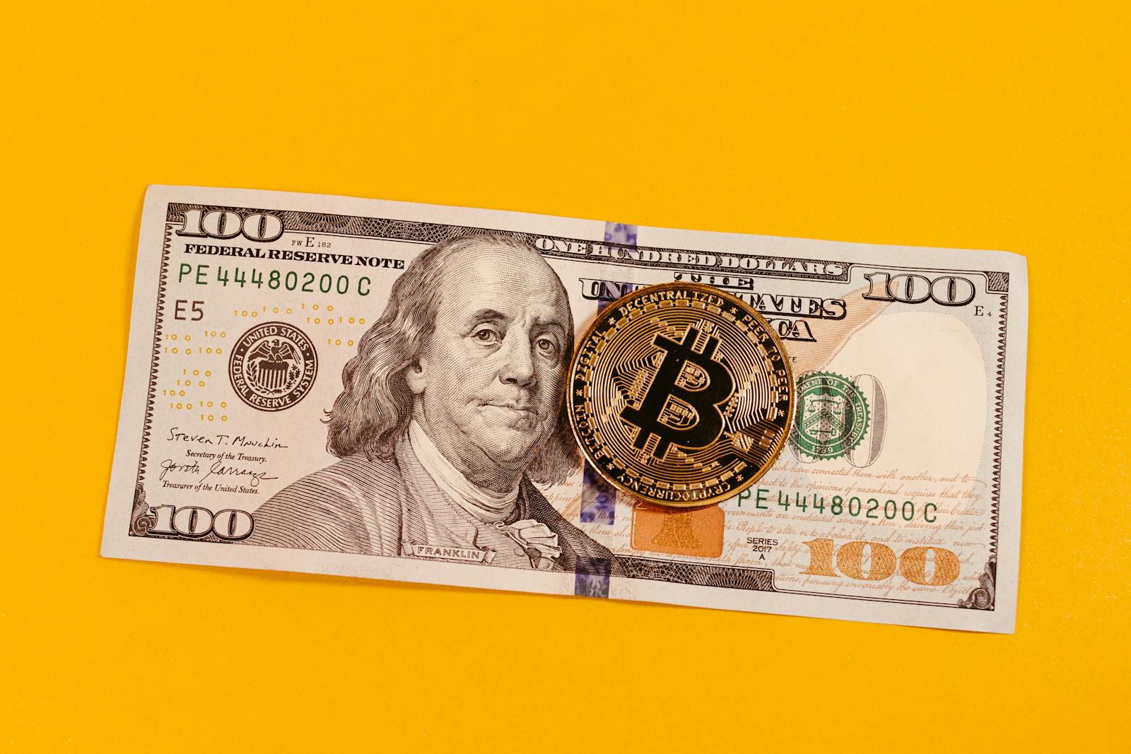 Gold bitcoin placed on a 100 US dollar bill over a bright yellow backdrop, symbolizing modern digital currency.