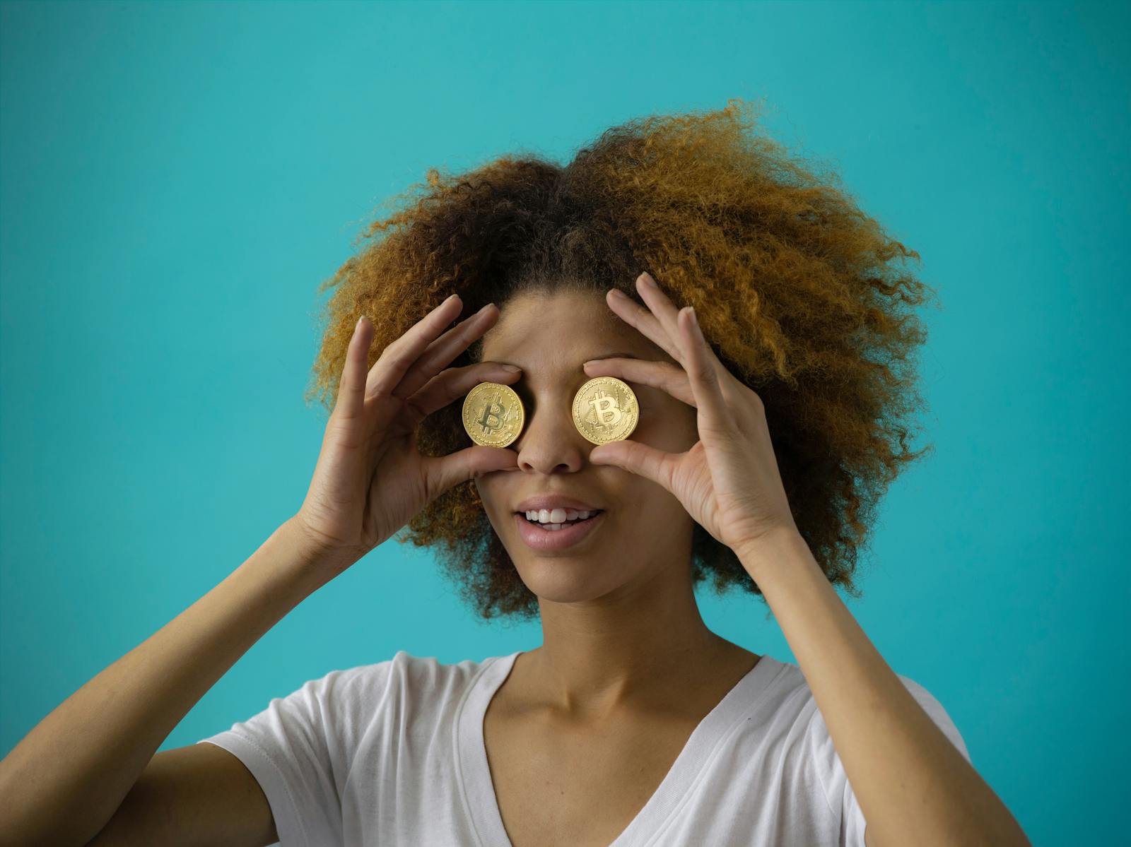 Smiling woman with afro hair holding cryptocurrency coins in front of eyes, against blue background.