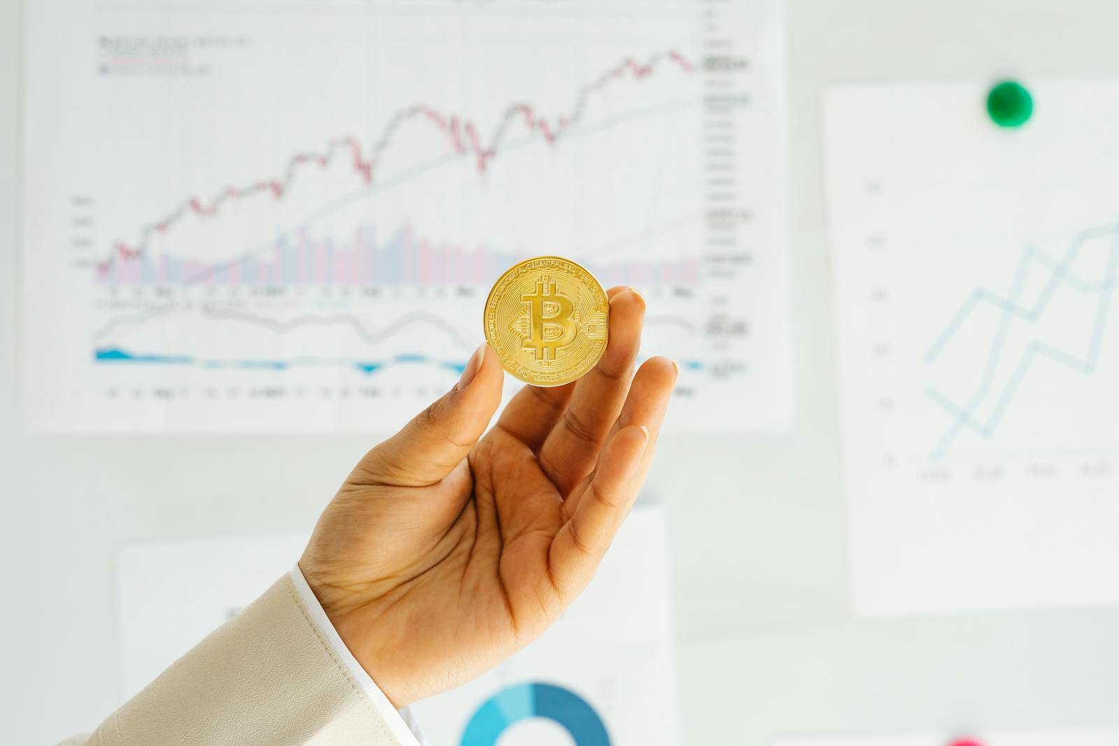 A hand holding a Bitcoin coin against financial charts depicting market trends.